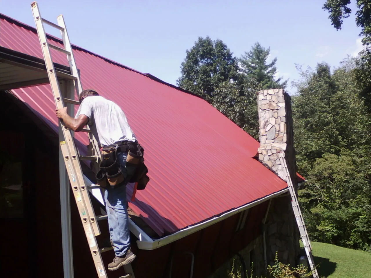 Roofer on a ladder beside a red metal roof and stone chimney — Soffit & Fascia Repair by Roofing Brothers in Continental